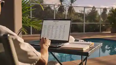 Person working through numbers on a laptop at a poolside table beside a screened pool enclosure
